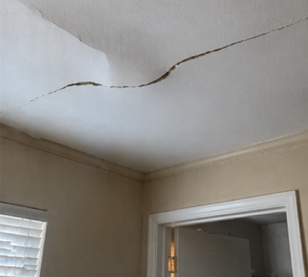 Interior ceiling with visible cracks and slight bowing beneath a sagging roof section.